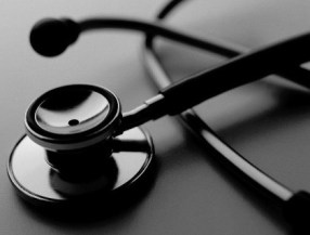 A black and white image showing a stethoscope on a table. The image shows the end of the stethoscope up close. 