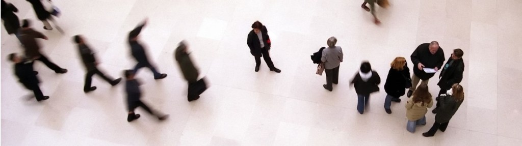 A group of adults standing at a museum. The photo is taken from overhead looking down on them. The person in the center is clear, while the others are blurred around them, as if they are moving. 